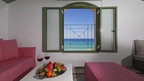 A room with a balcony, a table with fruit and wine glasses, and a view of the sea.