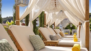 A row of sun loungers with white cushions and patterned pillows, arranged under a canopy with white drapes.