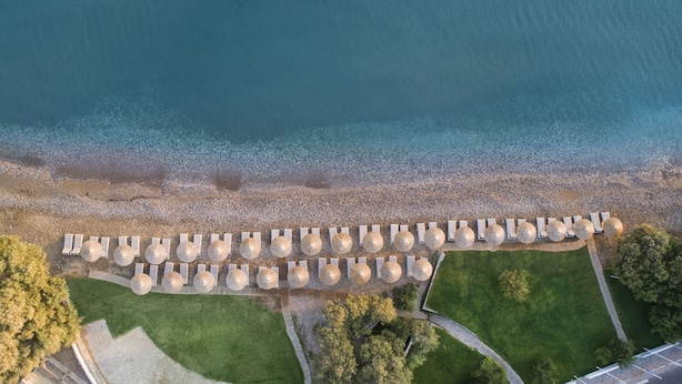 An aerial view of a beachside resort with rows of sun loungers and umbrellas, a pebble beach, and green lawns.