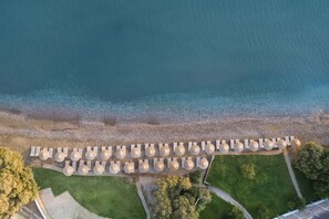 An aerial view of a beachside resort with rows of sun loungers and umbrellas, a pebble beach, and green lawns.