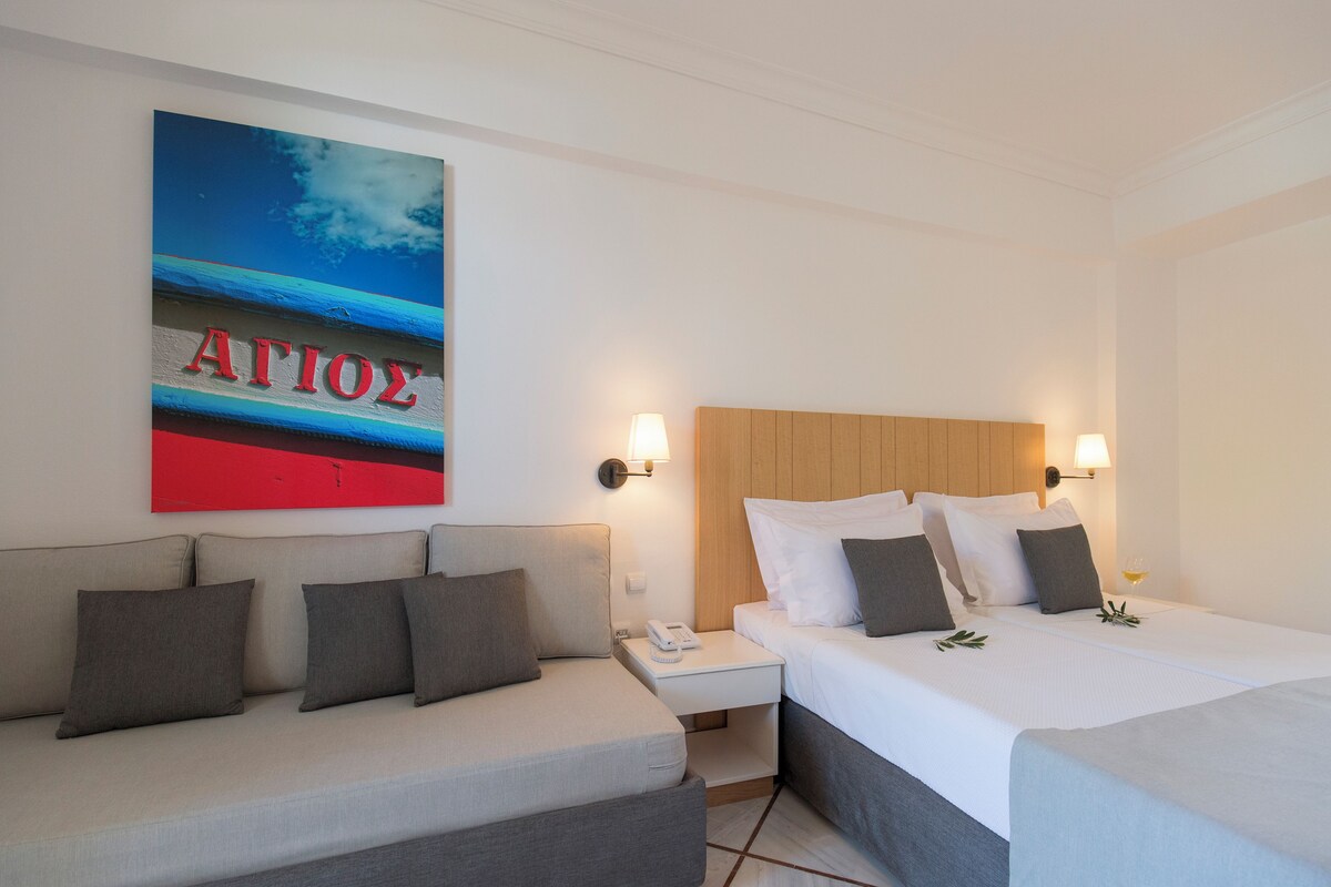 A hotel room with a bed, a sofa, a nightstand with a phone, and a wall with a painting.