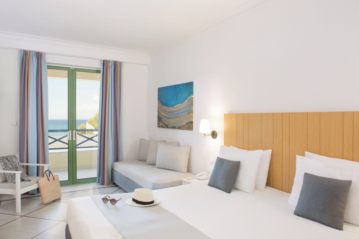 A hotel room with a bed, a sofa, a desk, and a view of the sea through a glass door.