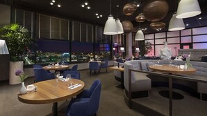 A modern restaurant with round tables and blue chairs, large hanging lights, and a city view through large windows. - Westcord WTC Hotel Leeuwarden (Leeuwarden)