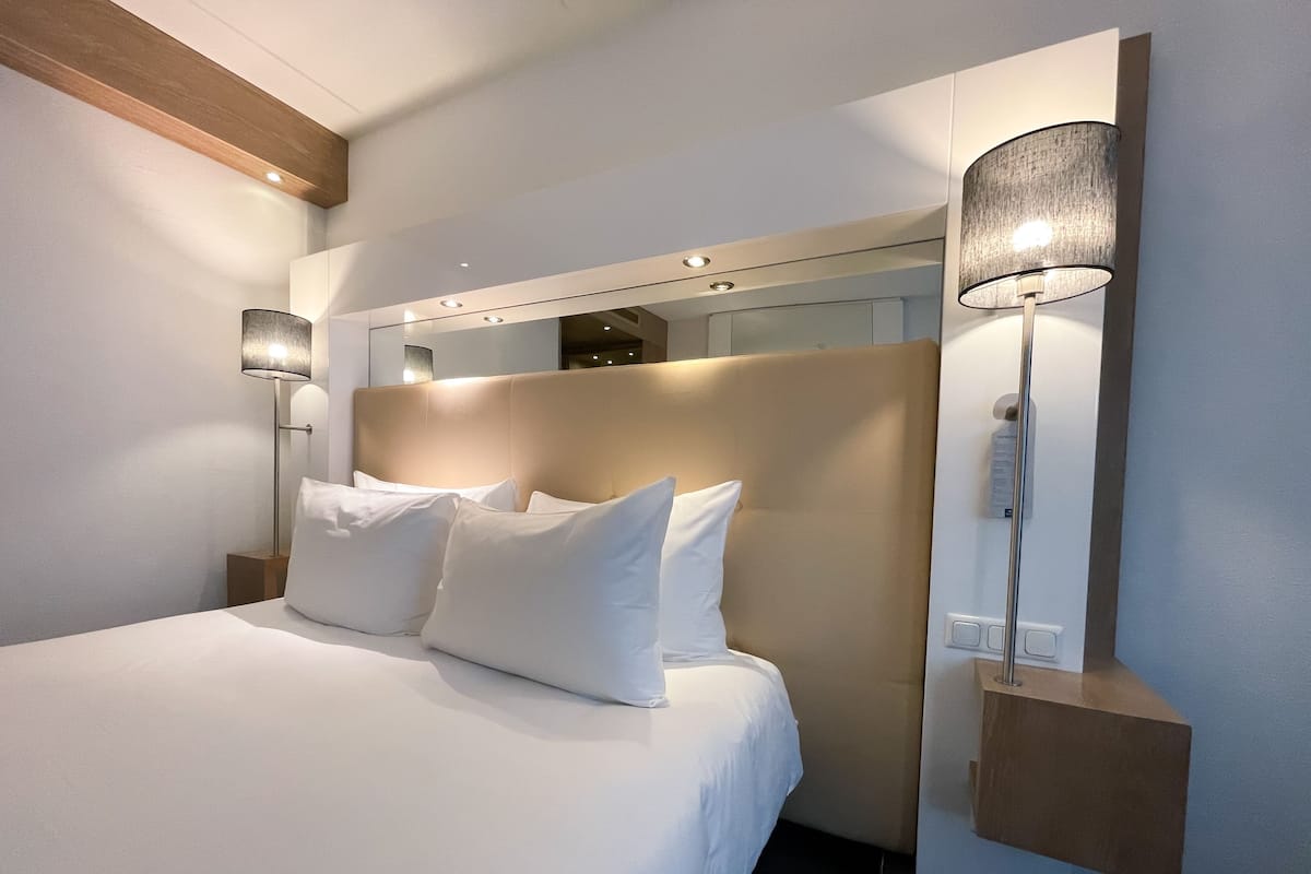 A modern hotel room with a large bed, a headboard, a bedside lamp, and a wall-mounted mirror.