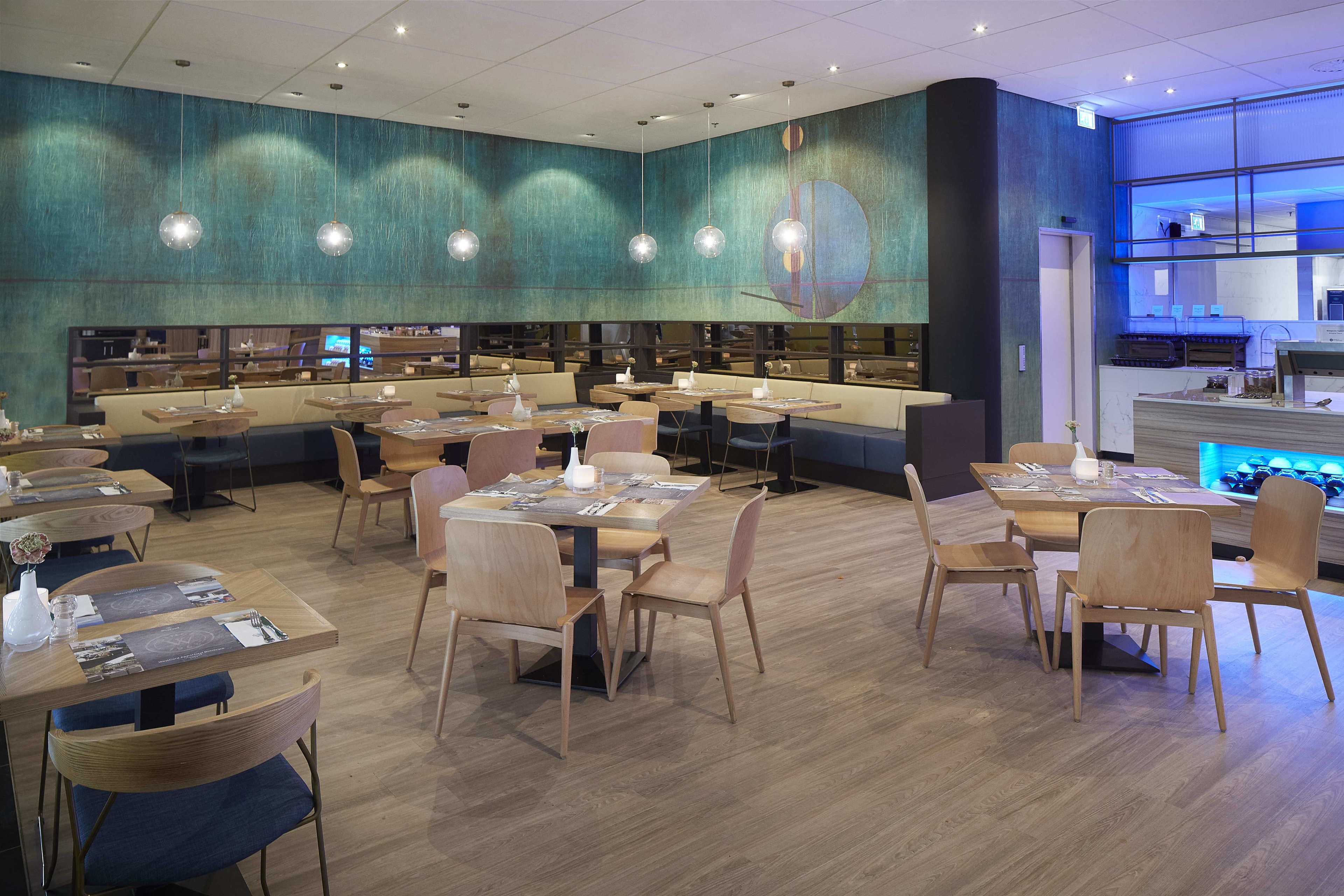 En moderne restaurant med flere spiseborde og stole, et stort vægmaleri og et køkkenområde, der kan ses gennem en glasvæg.
