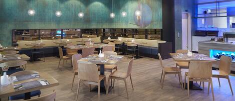 Un restaurante moderno con varias mesas y sillas, un mural grande en la pared y una cocina visible a través de un separador de vidrio.