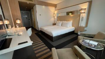 Habitación de hotel con cama, mesita de noche, silla, escritorio, lámpara y espejo.