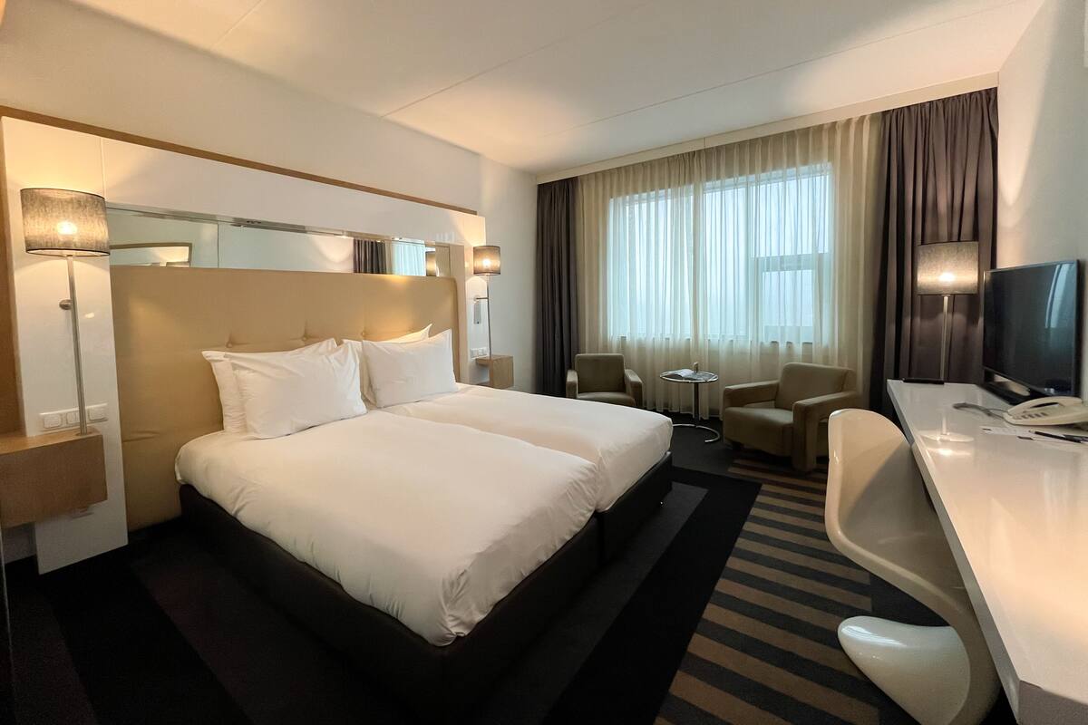 A hotel room with a large bed, two bedside tables, a desk with a chair, a TV, and a window with curtains.