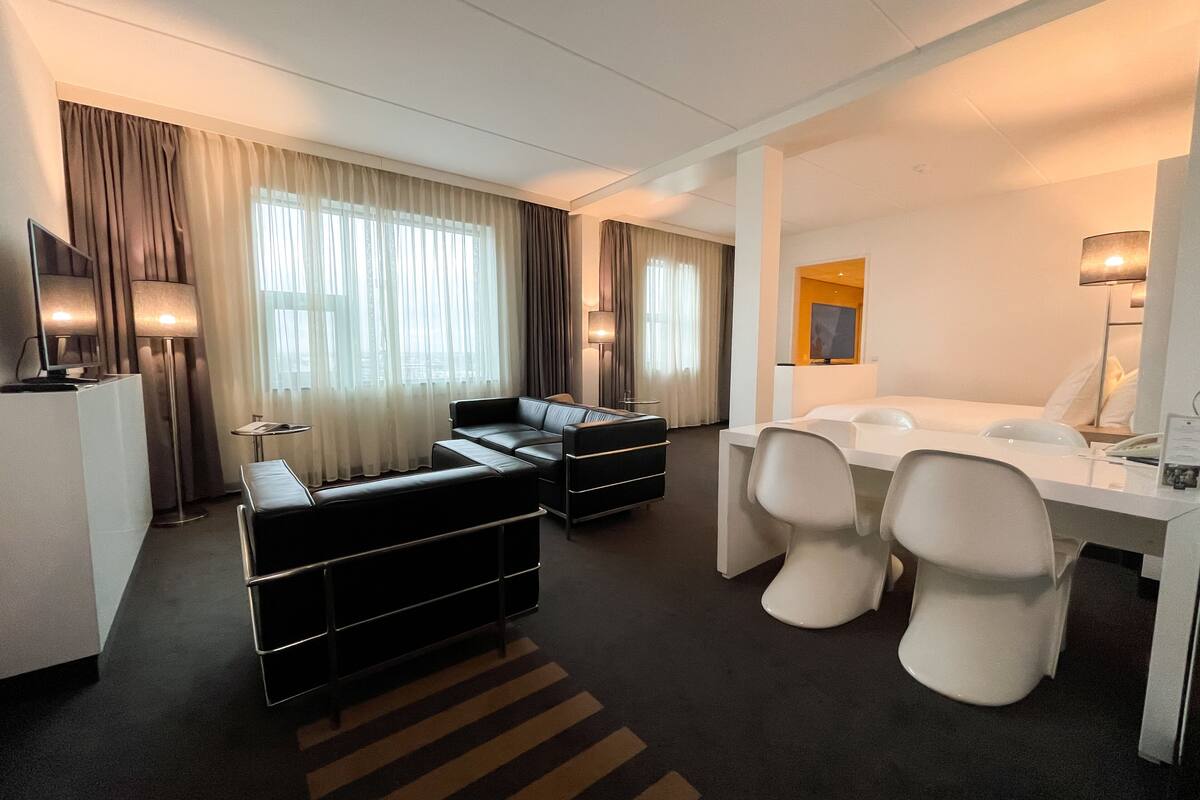 A modern hotel room with a sofa, a dining table with chairs, a TV, and a desk with a lamp.