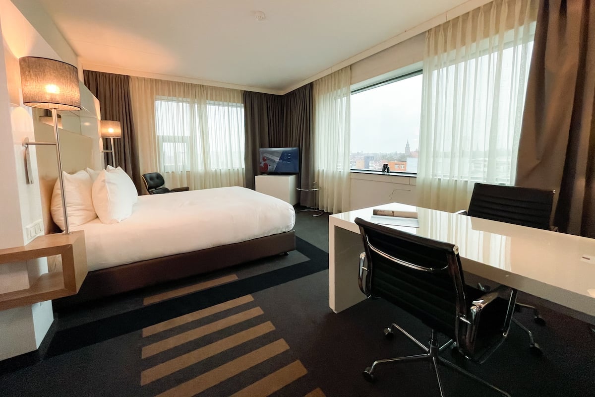 A hotel room with a bed, desk, chair, TV, and large windows with curtains.