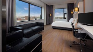 A modern hotel room with a large bed, a black leather sofa, a desk with a chair, and large windows offering a city view. - Westcord WTC Hotel Leeuwarden (Leeuwarden)
