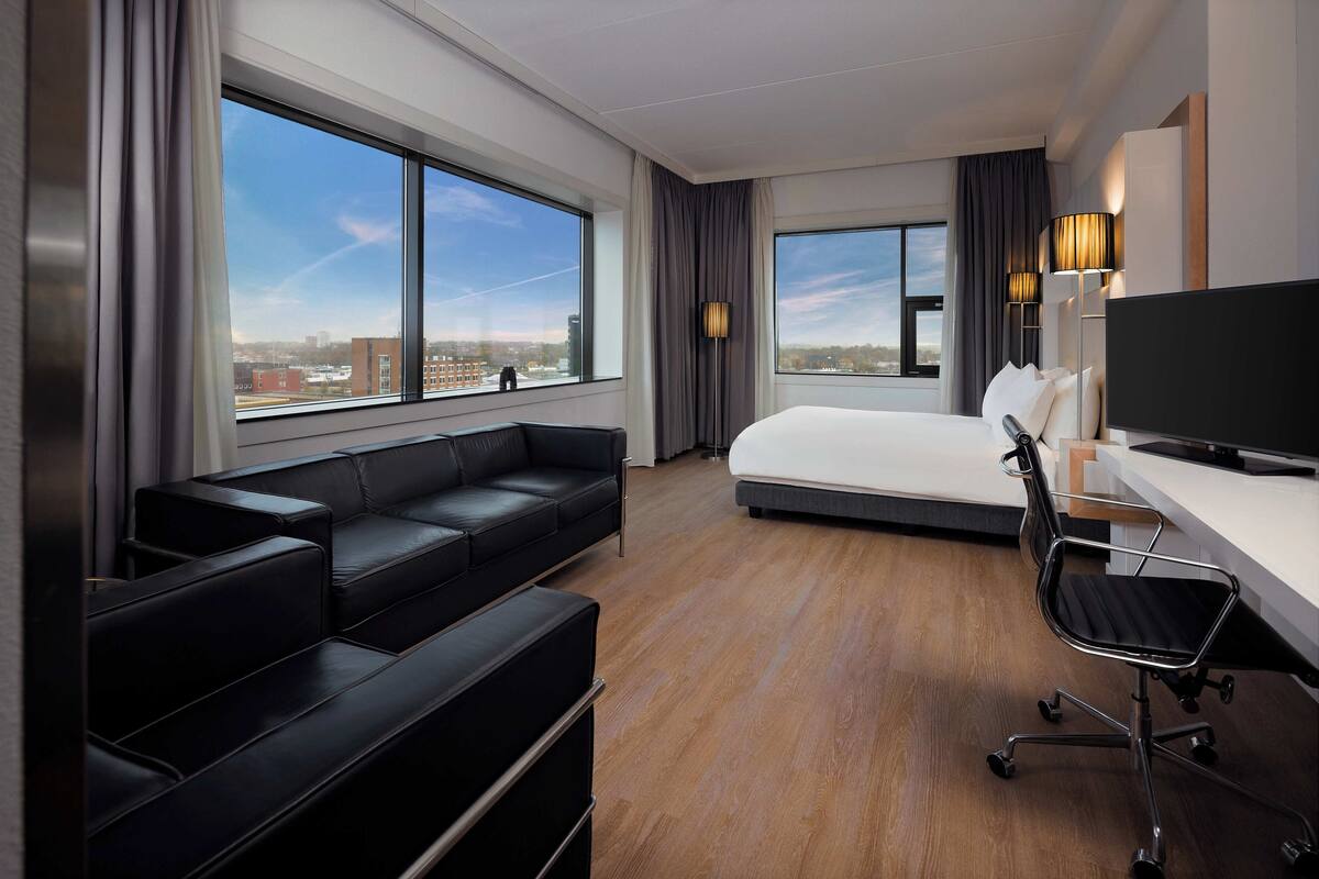 A modern hotel room with a large bed, a black leather sofa, a desk with a chair, and large windows offering a city view.