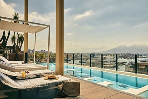 A rooftop infinity pool with lounge chairs and a view of a cityscape and a mountain.