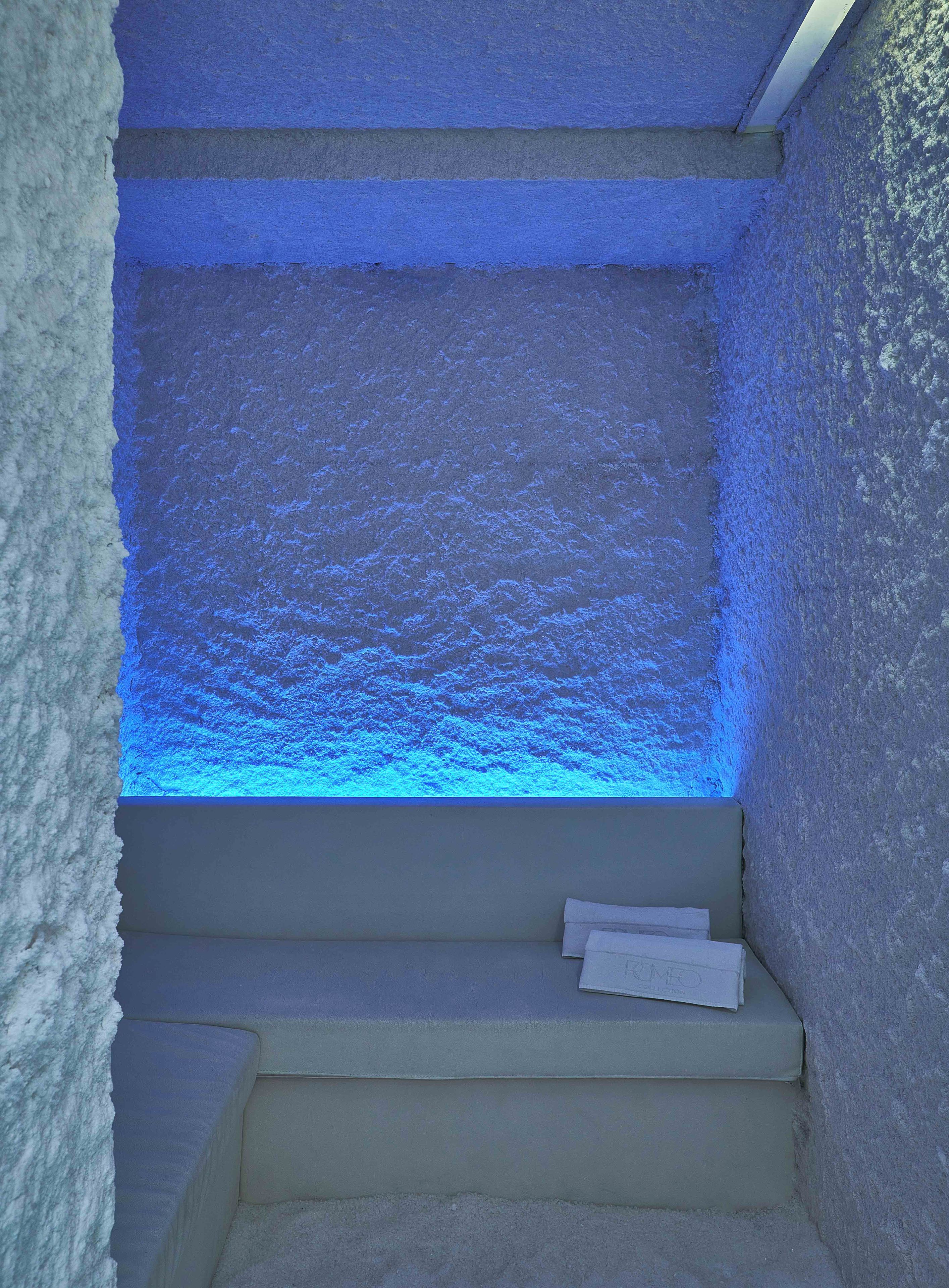 A room with a textured wall and a blue-lit window. There are steps leading to a bed with a white pillow and a white object on it.