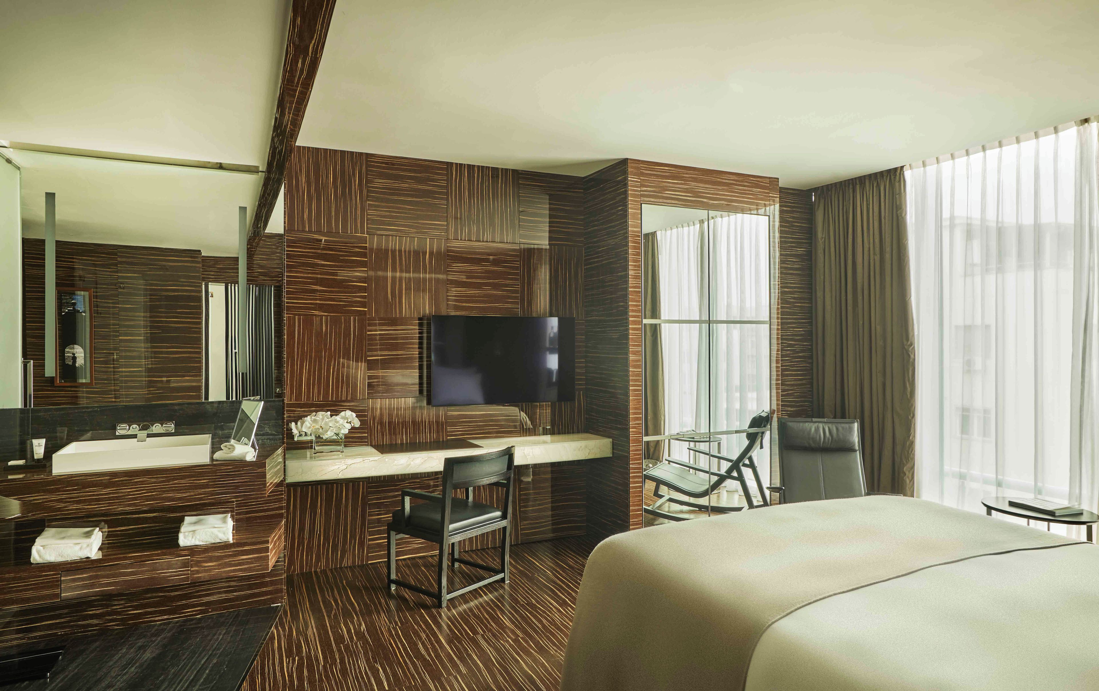 A hotel room with a large bed, a desk with a chair, a TV, a bathroom with a mirror, and a window with curtains.