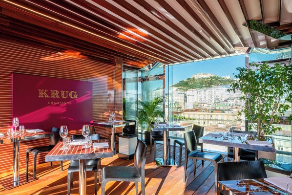 A modern restaurant with a view of a cityscape, featuring wooden flooring, glass tables, and a prominent Krug Champagne sign.