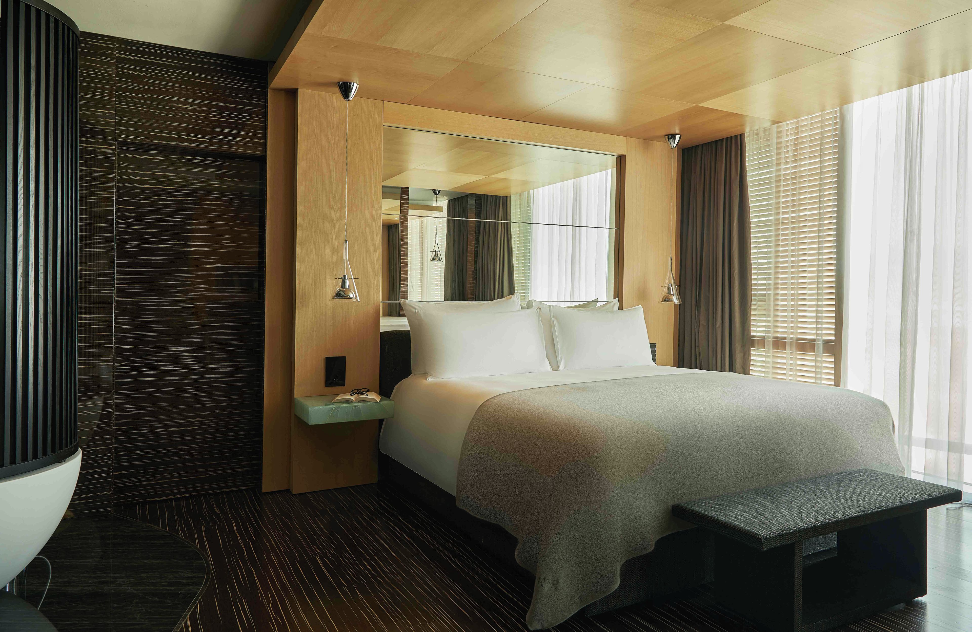 Una habitación de hotel moderna con una cama grande, una mesita de noche con un libro, un banco y un amplio ventanal con persianas.