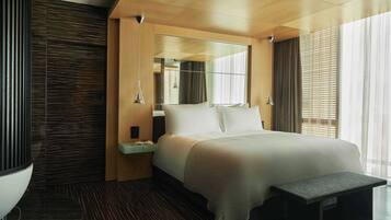 Una habitación de hotel moderna con una cama grande, una mesita de noche con un libro, un banco y un amplio ventanal con persianas.