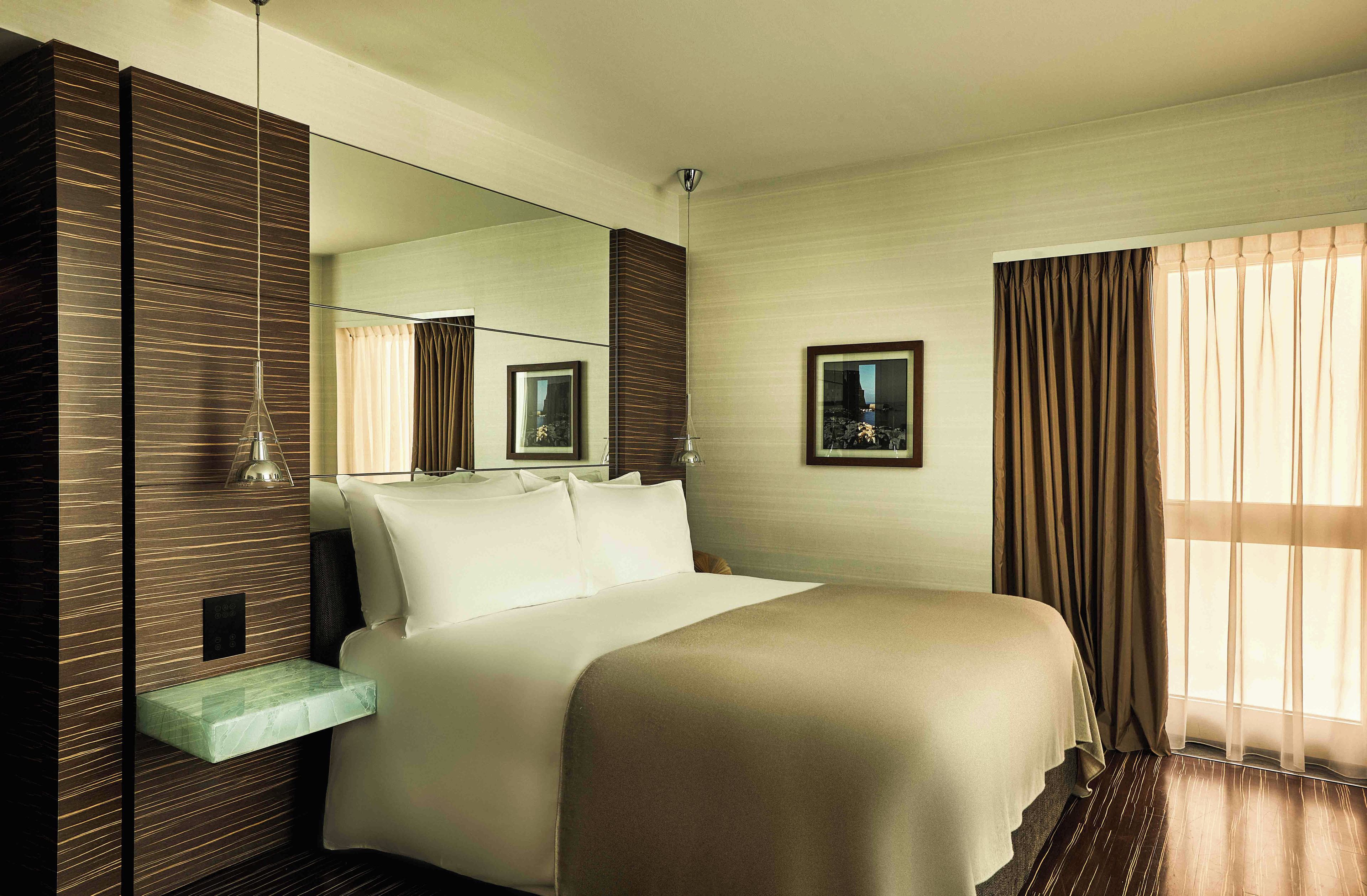 A hotel room with a bed, a bedside table, a mirror, a picture frame, and a window with curtains.