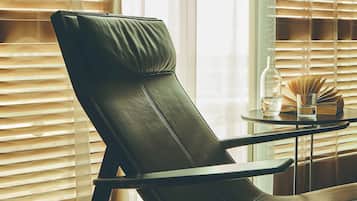 Una habitación con un sillón de cuero negro, una mesita redonda con una botella y un vaso, y una ventana con persianas.