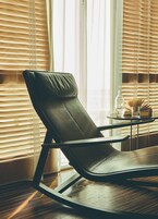 Una habitación con un sillón de cuero negro, una mesita redonda con una botella y un vaso, y una ventana con persianas.