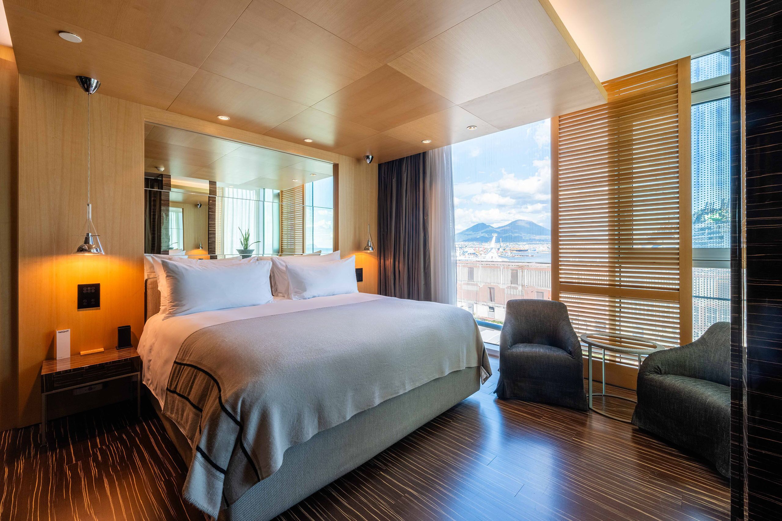Una habitación de hotel moderna con una cama grande, dos sillones, una mesita y un ventanal con persianas.