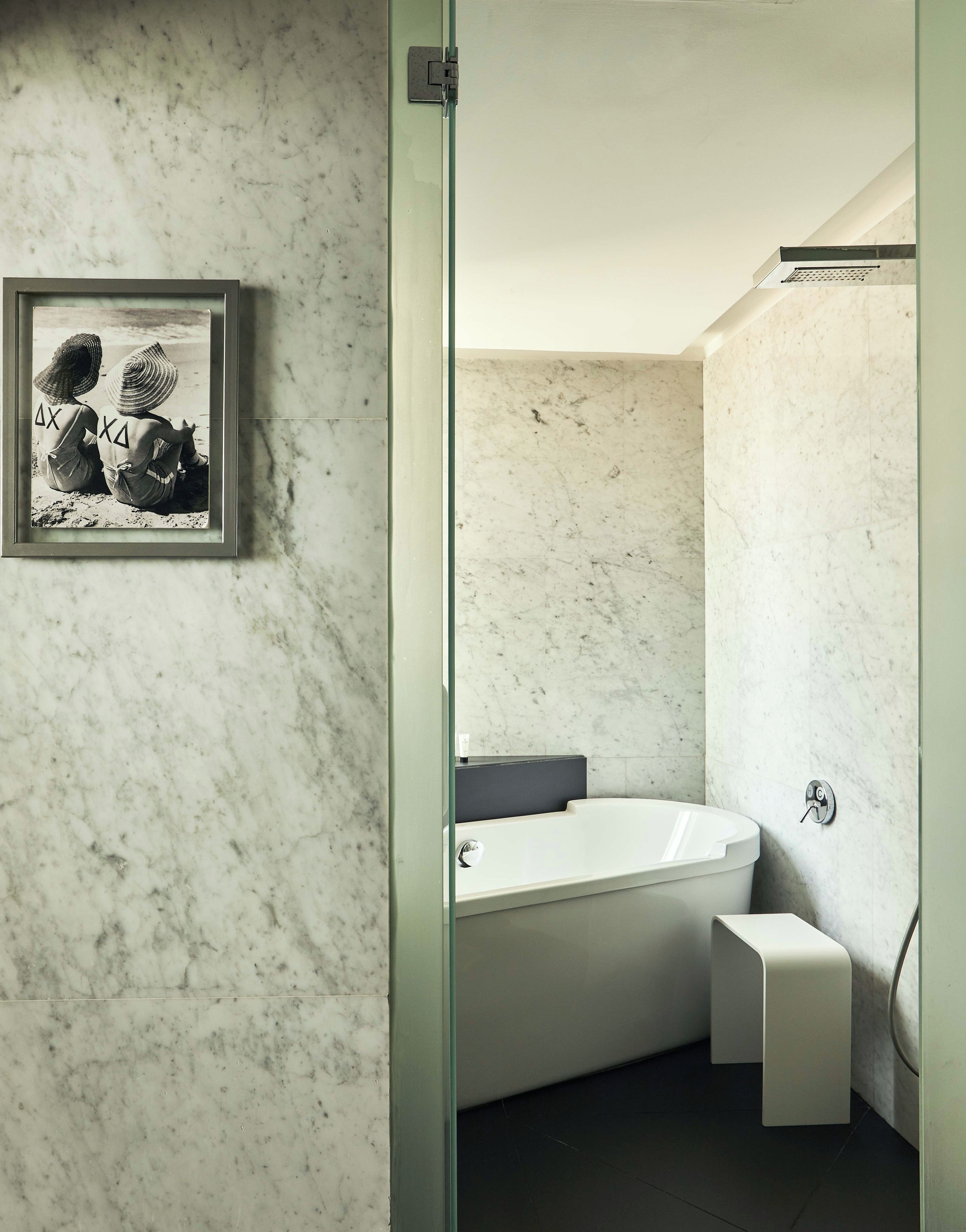 Un baño con pared de mármol, una fotografía enmarco, una bañera y un taburete pequeño.
