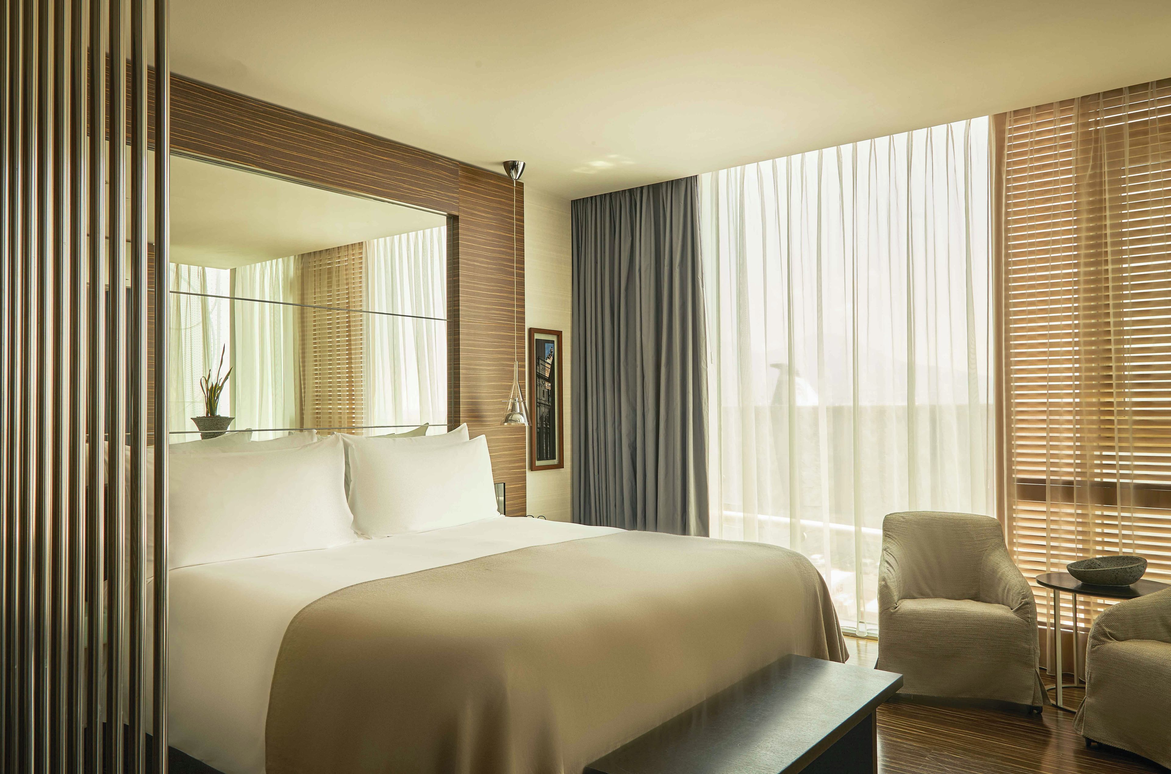 Une chambre d’hôtel avec un grand lit, une chaise, une petite table et une fenêtre avec des stores.