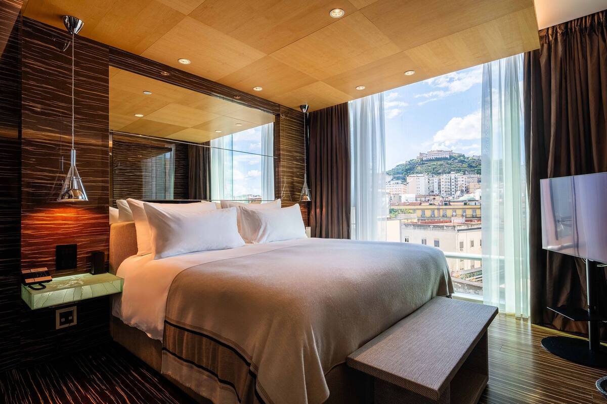 A modern hotel room with a large bed, a bedside table, a TV, and a view of the city.