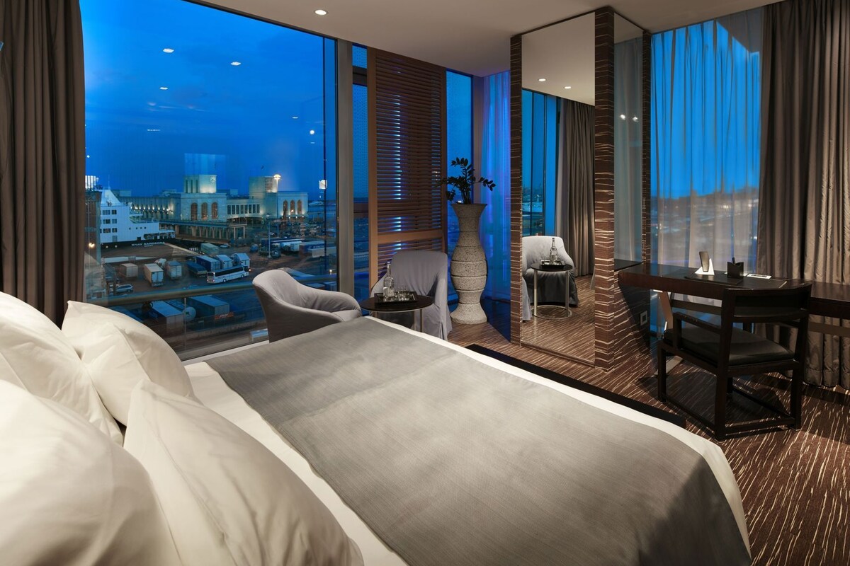 A modern hotel room with a large bed, a desk, a chair, and a view of the cityscape at night.