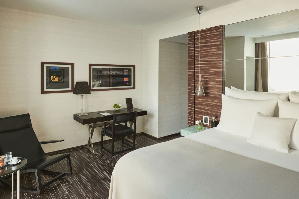 A hotel room with a bed, a desk with a chair, a small table with a cup and saucer, and a mirror on the wall.
