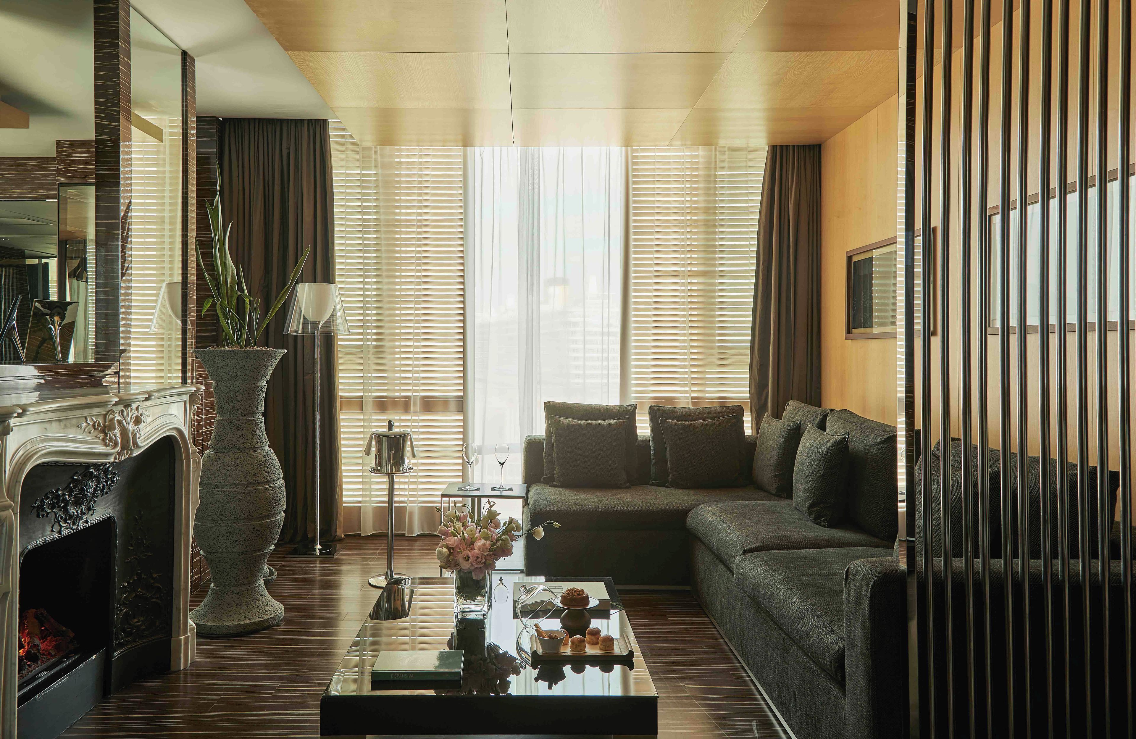 A modern living room with a fireplace, a sofa, a coffee table with decorative items, and a large window with blinds.