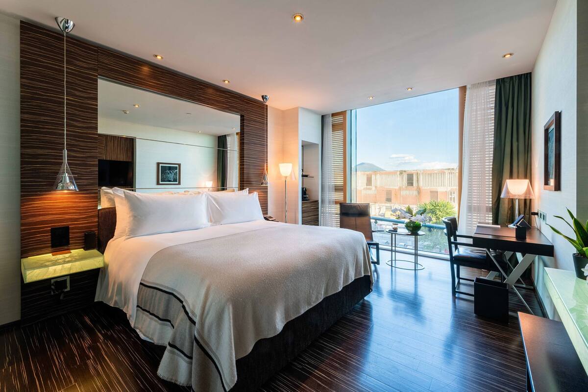 A modern hotel room with a large bed, a desk, and a balcony with a view.
