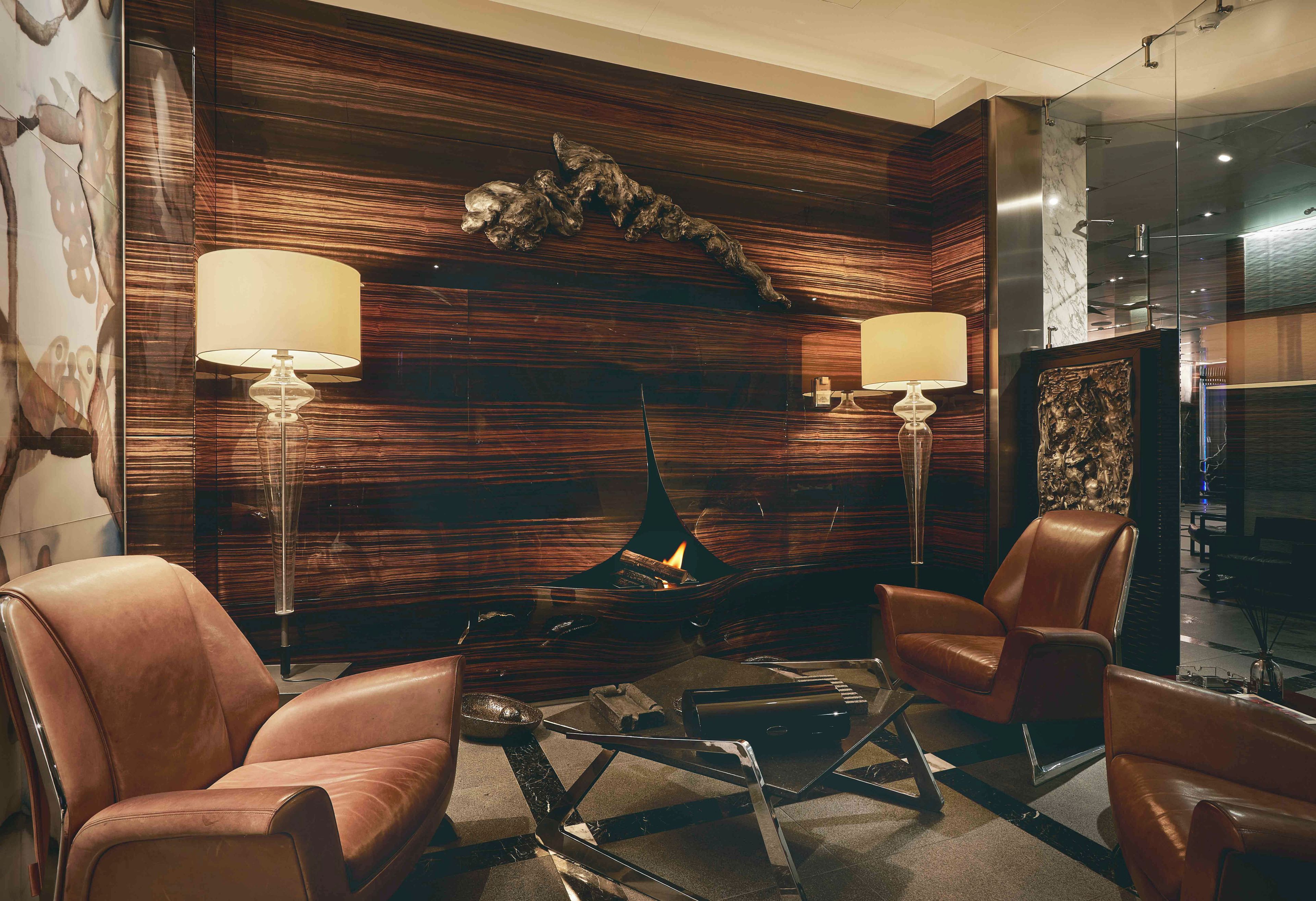 A modern lounge area with leather armchairs, a glass coffee table, and a fireplace with two lamps.
