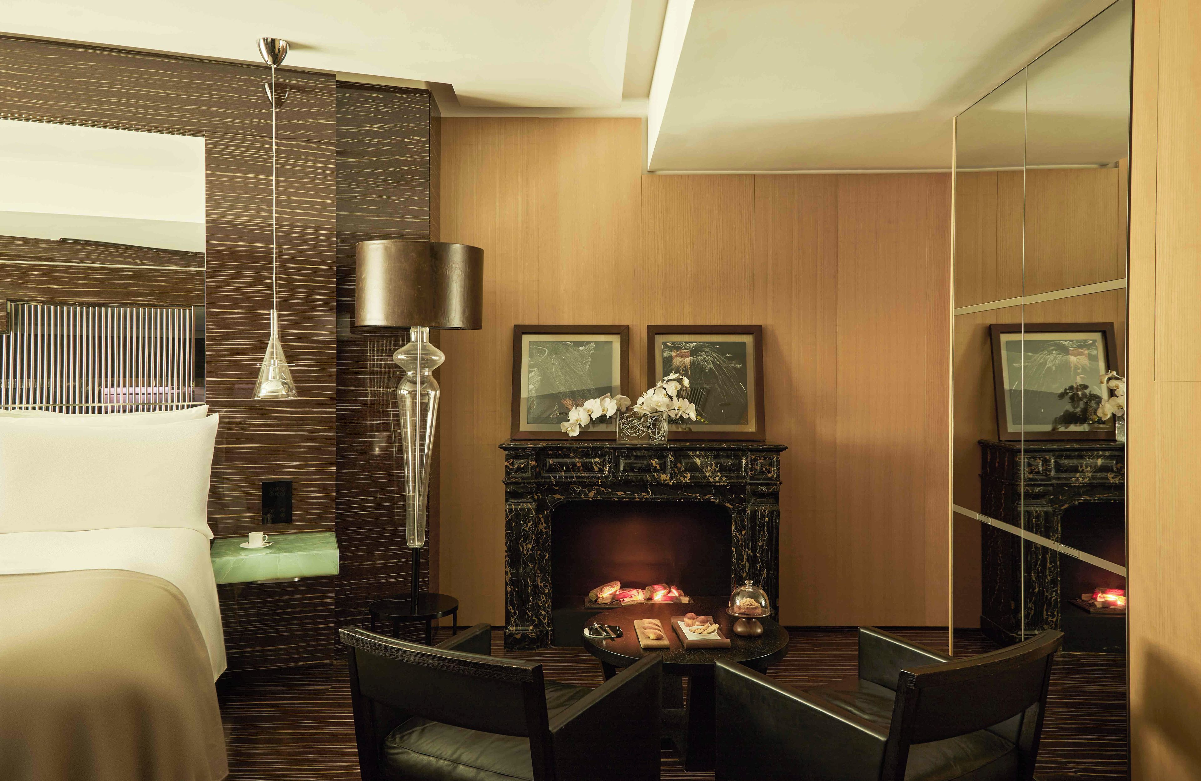 A modern hotel room with a bed, a fireplace, a dining table with chairs, and a large mirror.