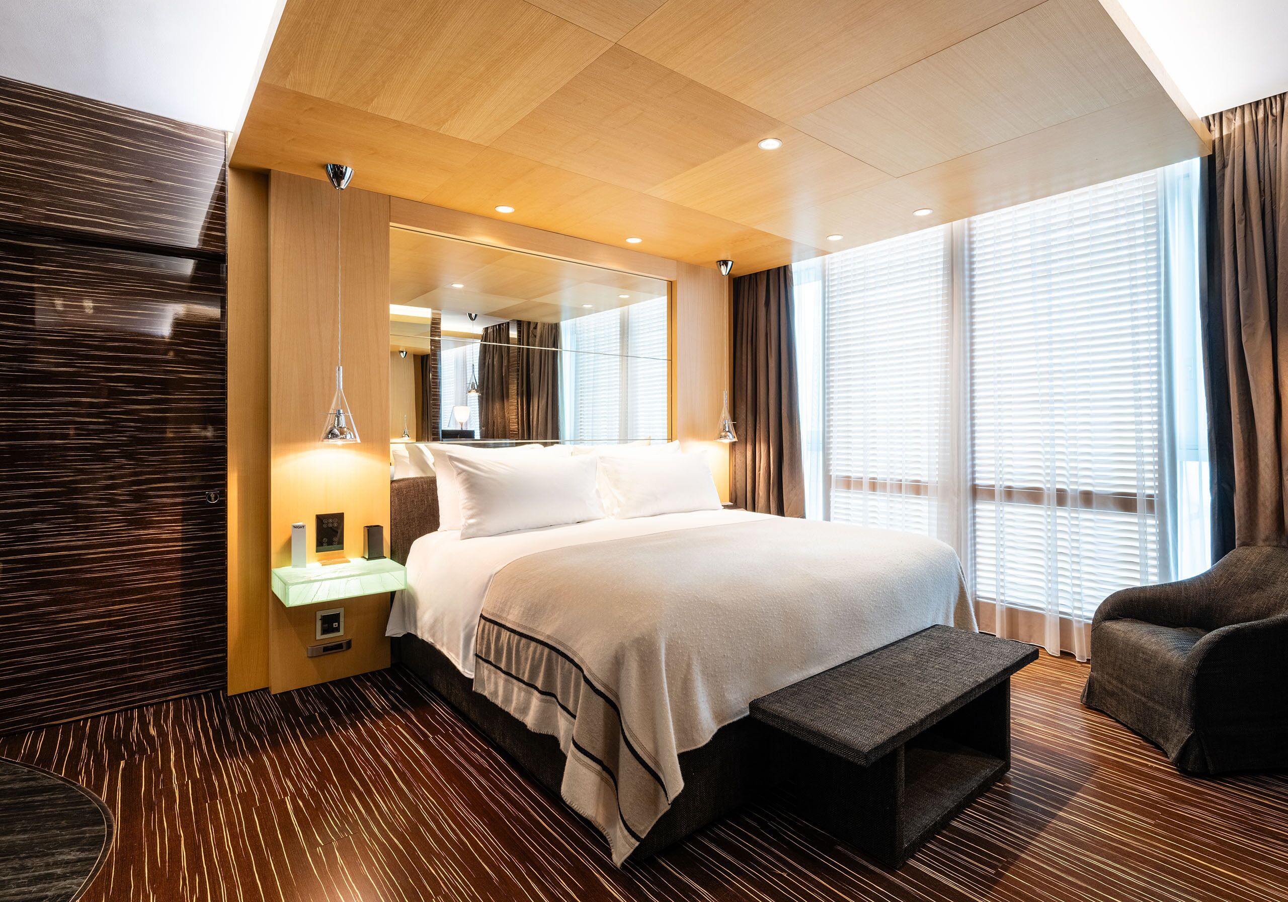 Une chambre d’hôtel avec un grand lit, des tables de chevet, un banc, un miroir et une vue sur l’extérieur grâce à de grandes fenêtres.