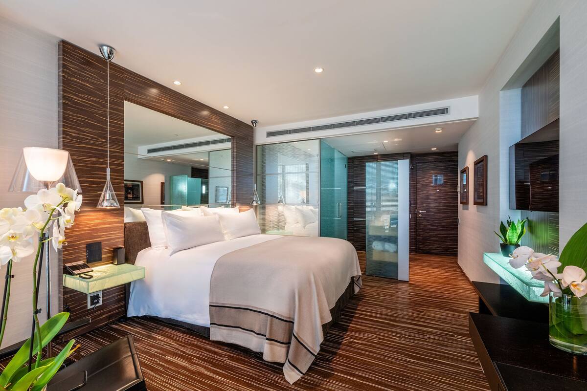 A modern hotel room with a large bed, a glass desk, a mirror, and a view of the city through large windows.