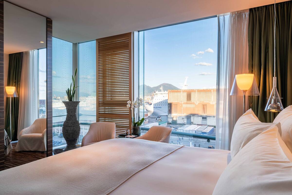 A hotel room with a large bed, two chairs, a vase with flowers, and a view of the cityscape through large windows.