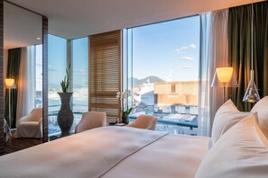 A hotel room with a large bed, two chairs, a vase with flowers, and a view of the cityscape through large windows.