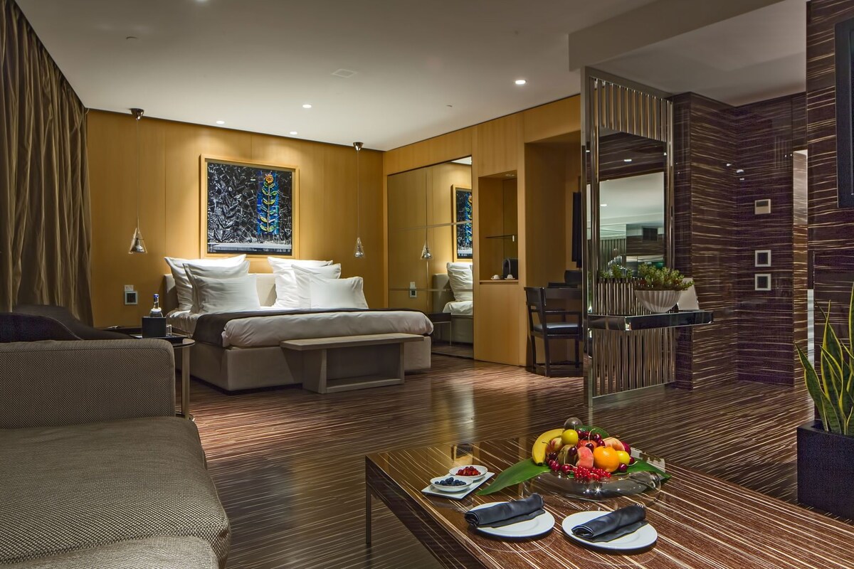 A modern hotel room with a bed, sofa, and dining area with fruit.