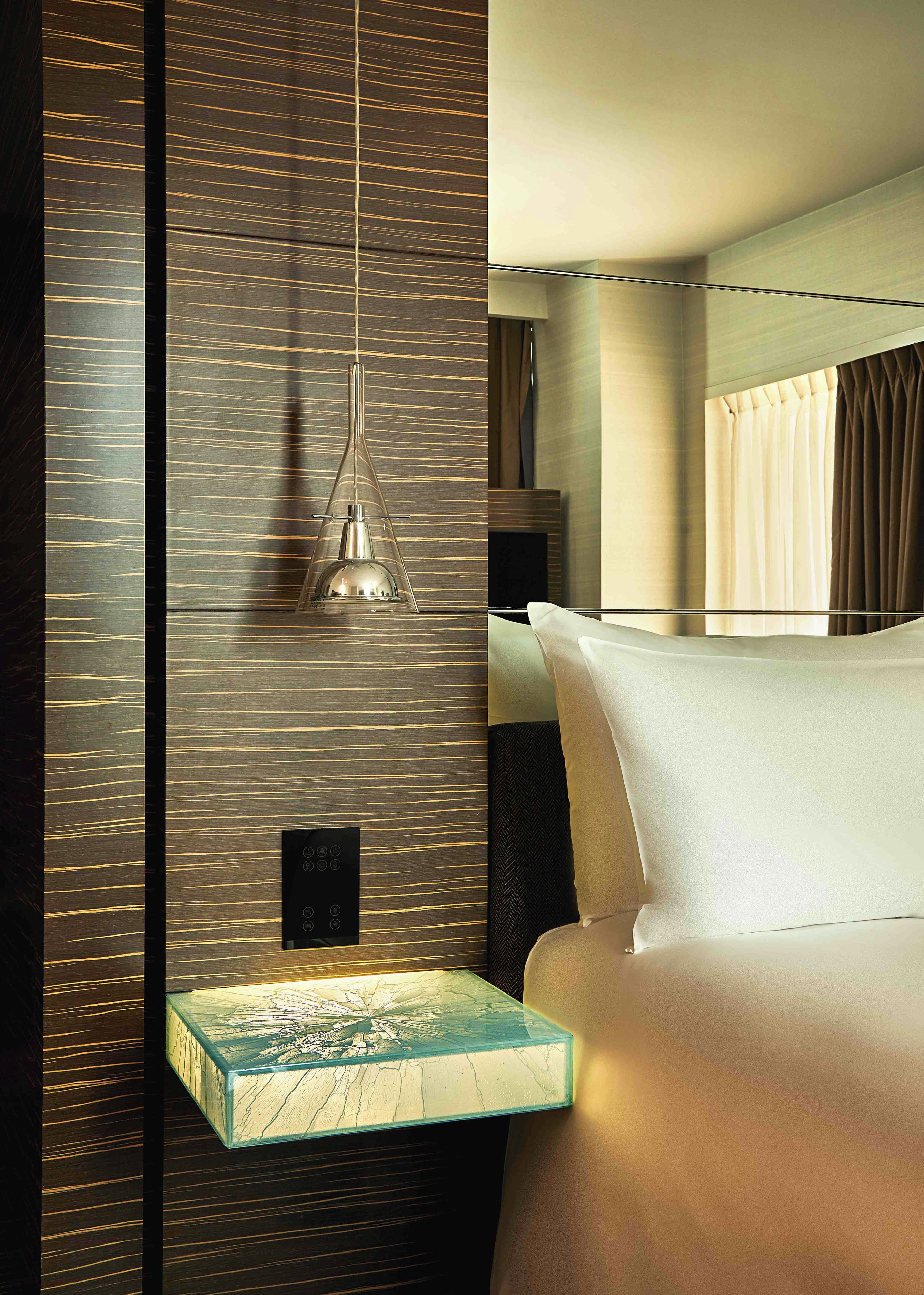 A modern hotel room with a bed, a bedside table with a glass top, a hanging pendant light, and a wall with a wood grain pattern.