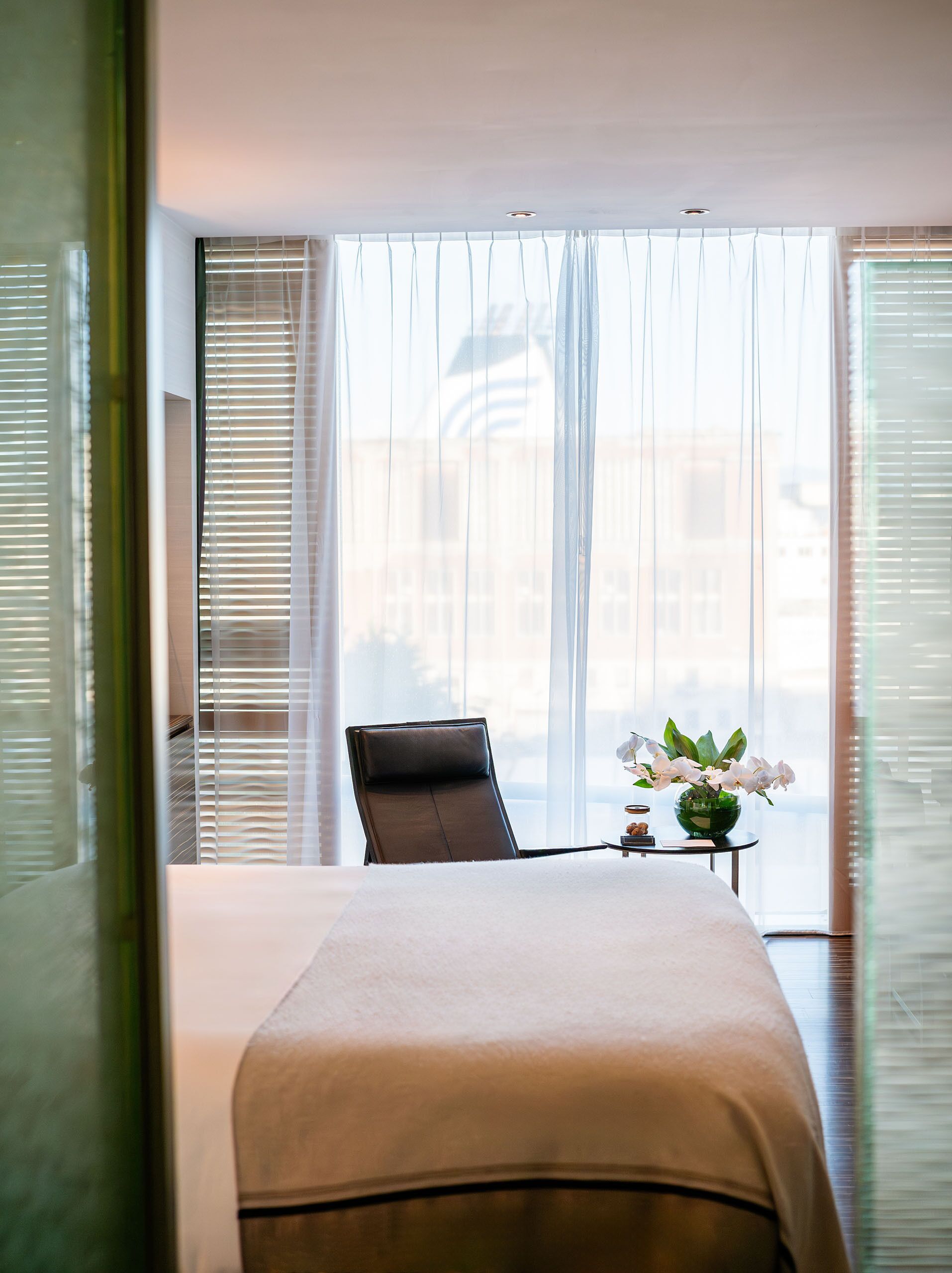 A hotel room with a bed, a chair, a small table with a vase of flowers, and large windows with blinds.