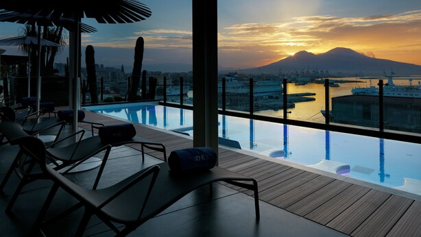 A rooftop infinity pool with lounge chairs and umbrellas, overlooking a cityscape and a mountain at sunset.