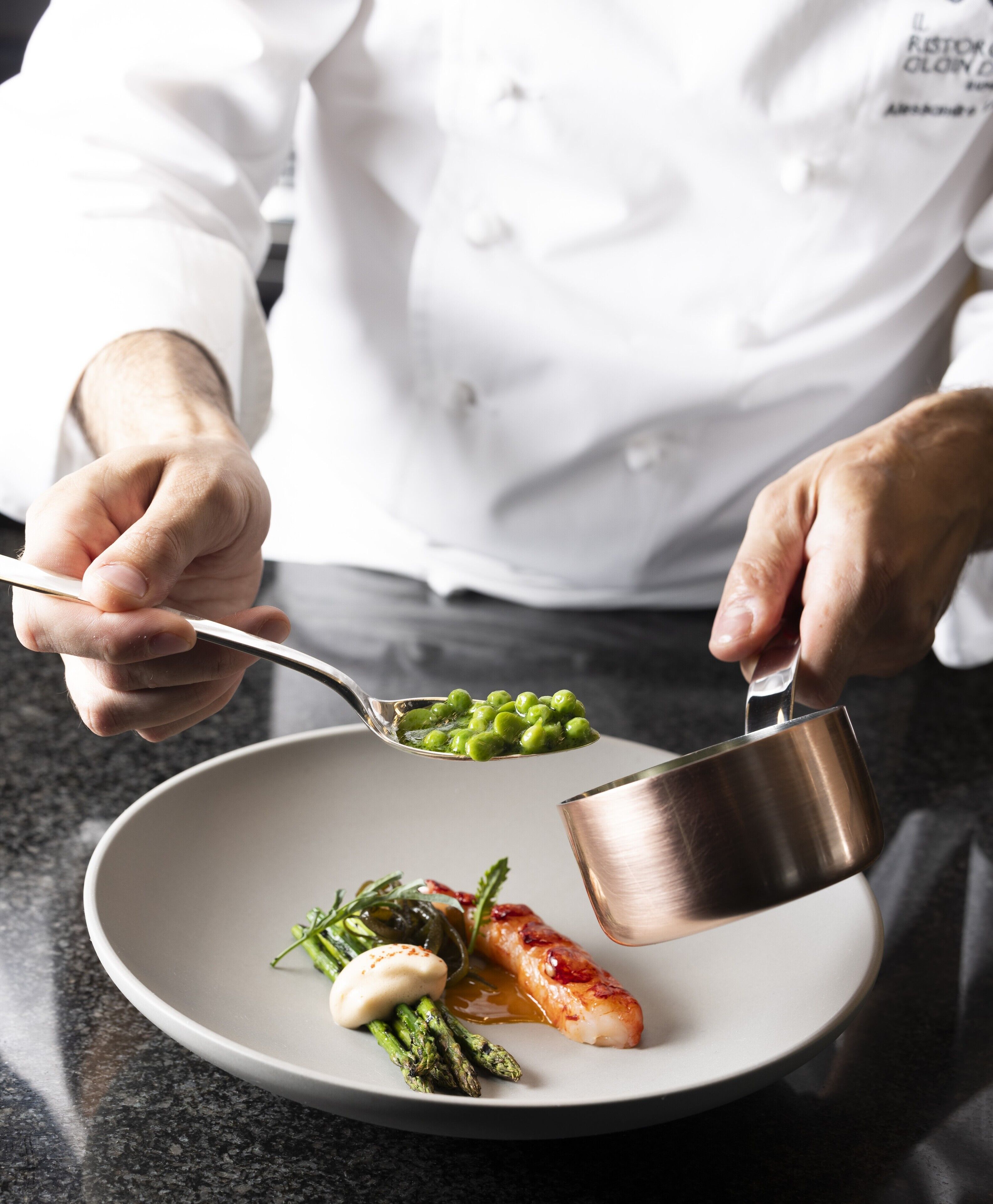 A chef holding a spoon with peas and pouring sauce from a metal cup onto a plate with asparagus and a piece of fish.