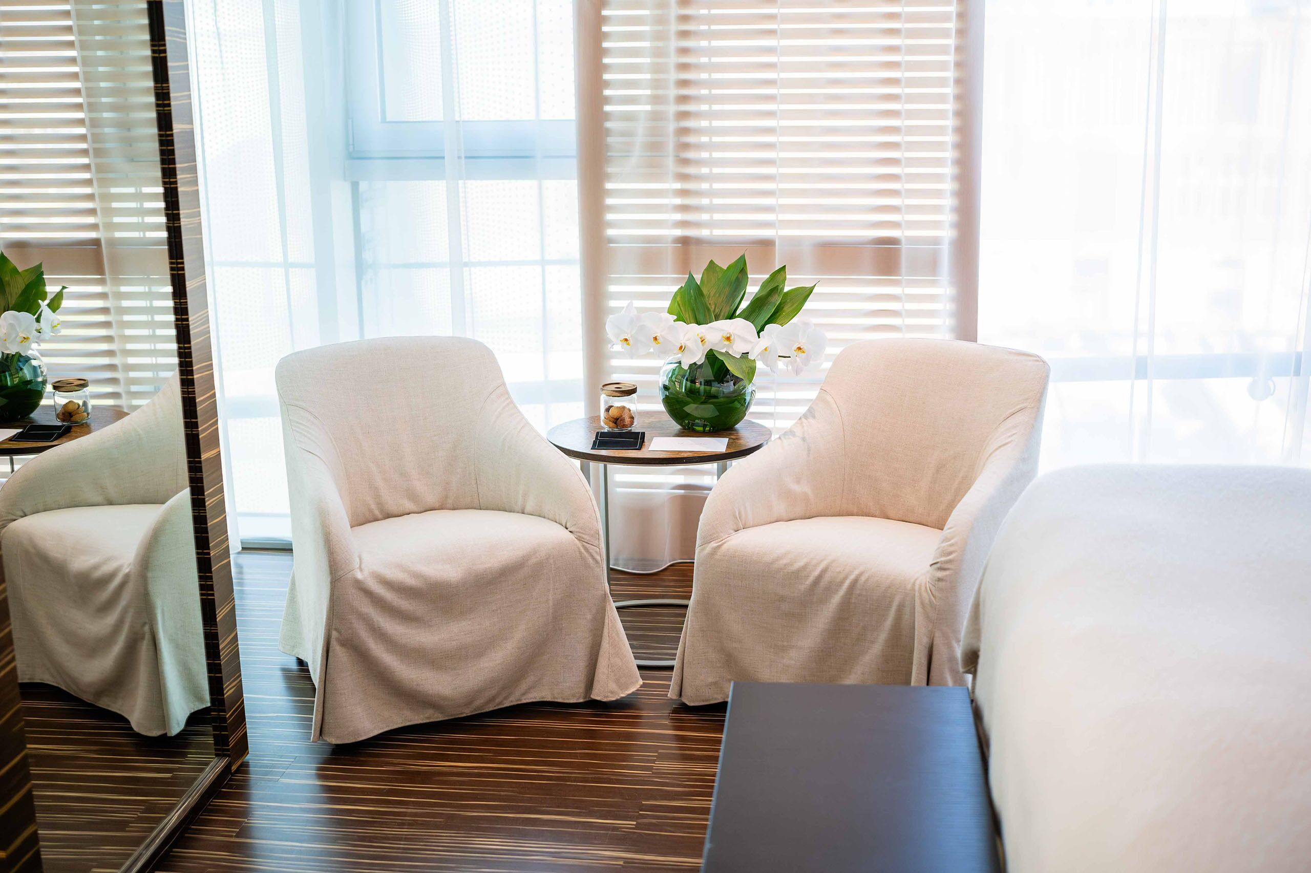 A hotel room with two beige armchairs, a small round table with a vase of white orchids, and a large mirror.