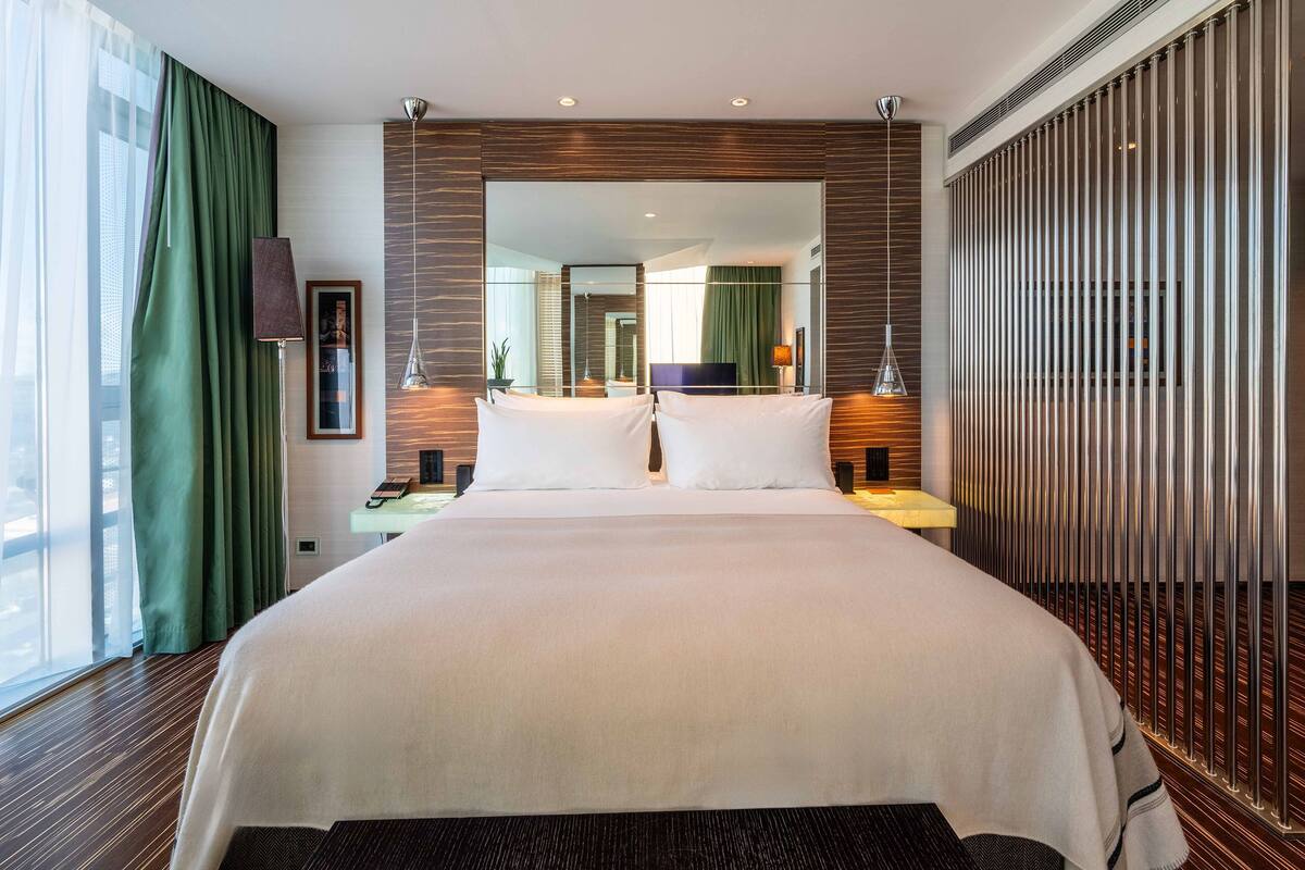 A large bed with white linens, a nightstand with a lamp, a large mirror, and a view of a cityscape through the window.