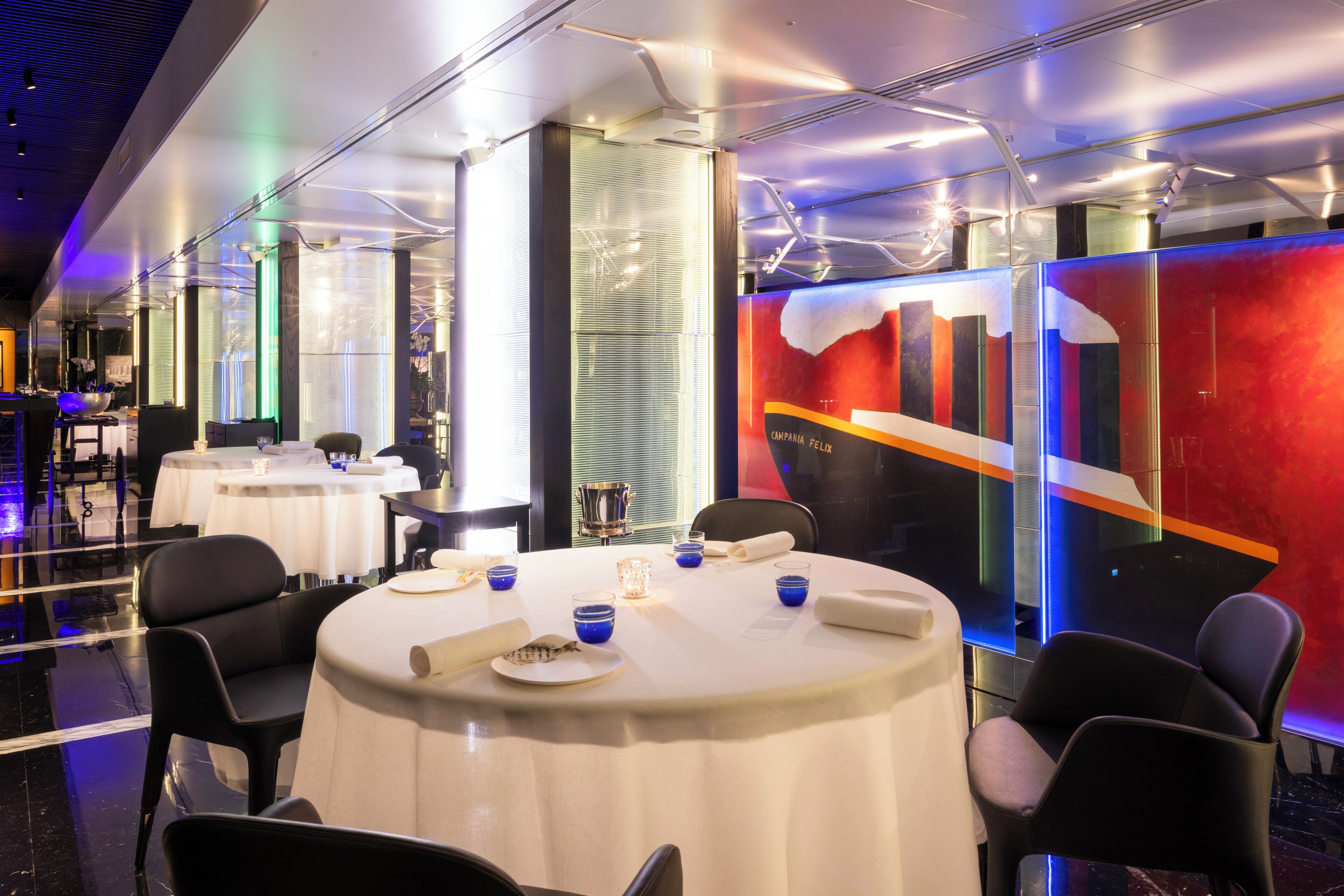 A modern restaurant with round tables, black chairs, and a colorful abstract mural on the wall.