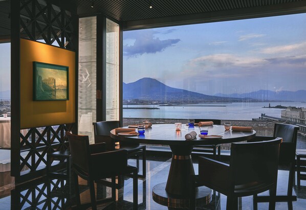 A dining area with a table set for four, overlooking a body of water and a mountain.