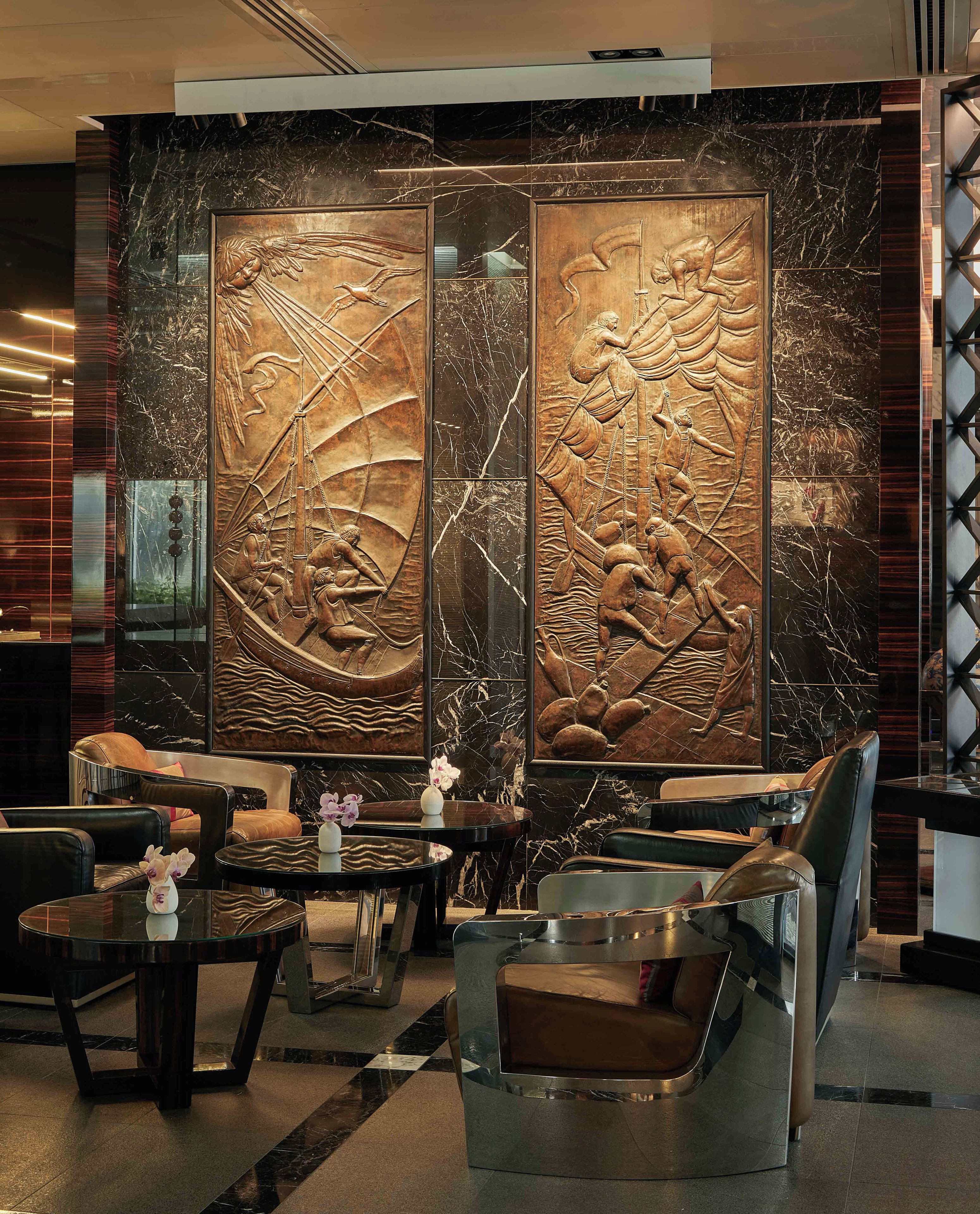 A lobby with a modern design, featuring a large decorative panel with intricate carvings, leather seating, and a glass coffee table with a vase of flowers.