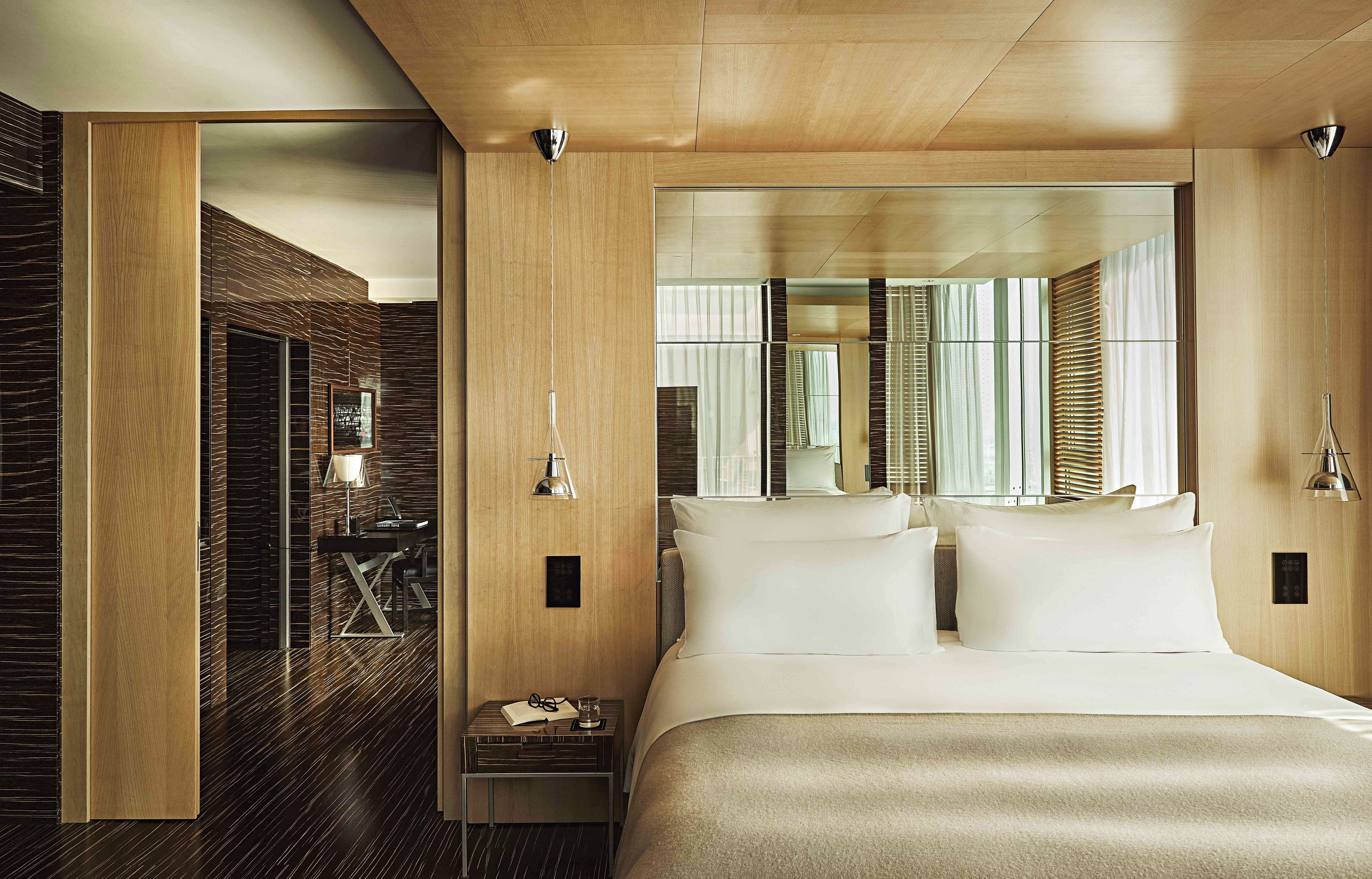 A modern hotel room with a large bed, a bedside table, a mirror, and a view of the city through the window.